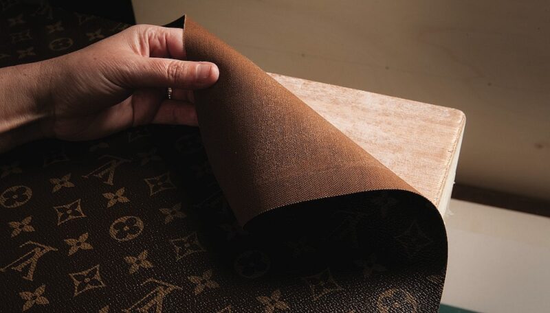 A person holds and lifts the corner of a textured, brown fabric with a distinctive pattern of logo2s, revealing a wooden surface underneath.